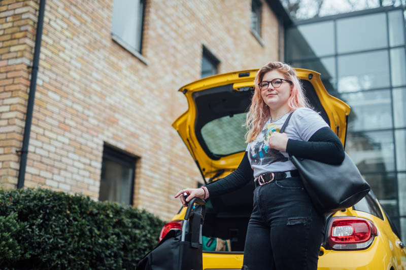 Image of Orla with car boot open, holding luggage trolley, and handbag