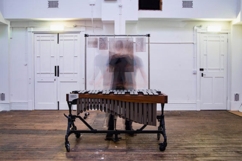 blurry, fast-moving person playing a vibraphone in an otherwise-empty, old, white-walled, dark-wooden-floored indoor space