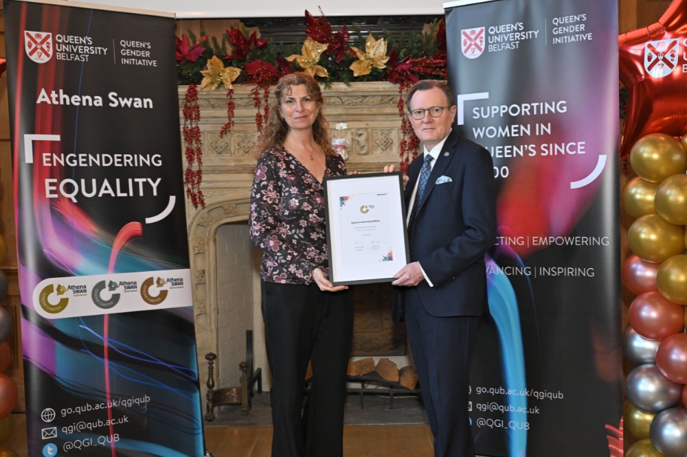 woman pictured with a man in a suit holding a framed award certificate in an old wood-paneled event hall with large fireplace and presentation screen in the background and pop-up stands and balloon pillars on either side