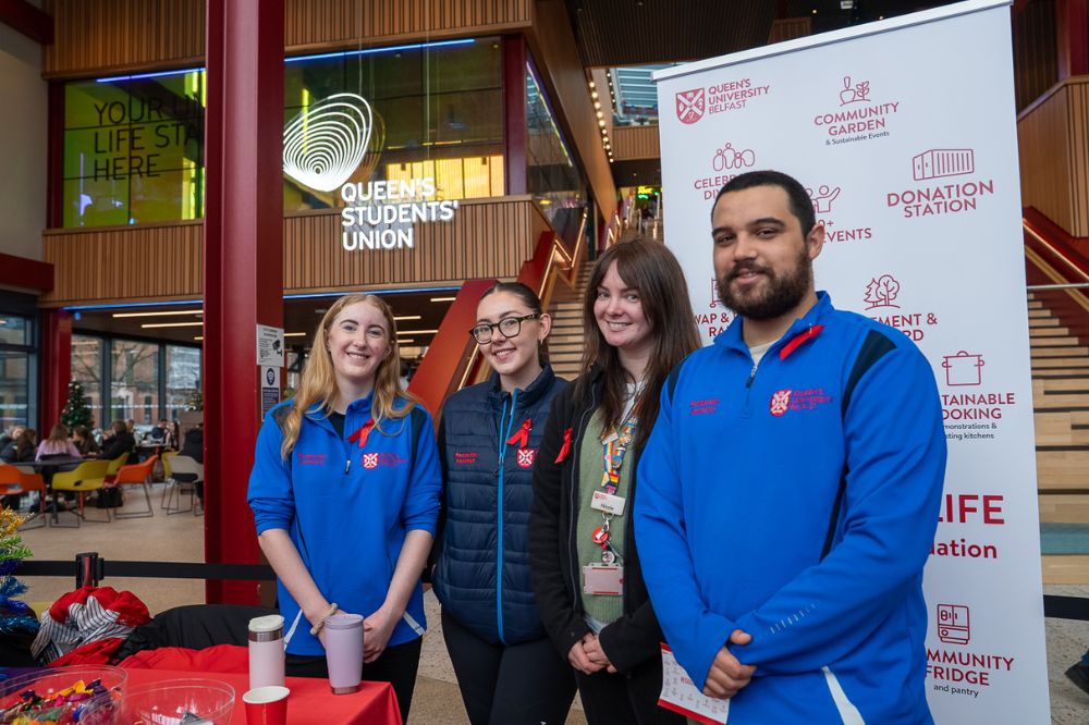 Four members of the Queen’s Students’ Union team stand smiling in front of a branded display inside the Students’ Union building.