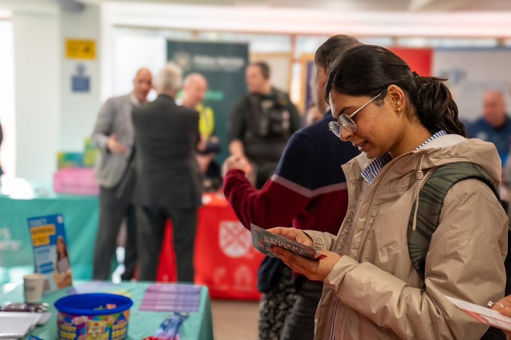 A student reads an information leaflet at an indoor campus event, with information stalls and attendees visible in the background.