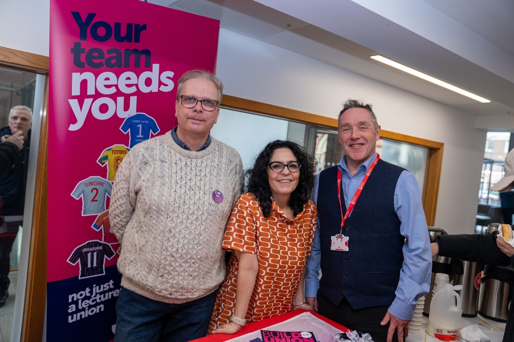 Three Staff Union members stand smiling at an indoor campus event beside a banner reading “Your team needs you