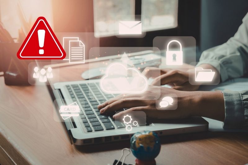 Person using laptop in a home-working scenario. Several less prominent data-themed icons surround the laptop with one prominent 'urgent' / 'beware' exclamation icon in the top left
