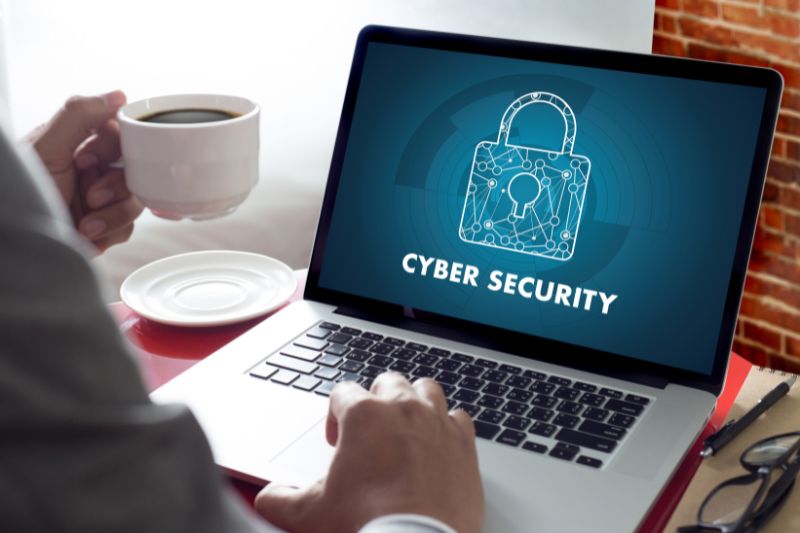 close-up view of person holding coffee sitting at laptop whose screen reads Cyber Security