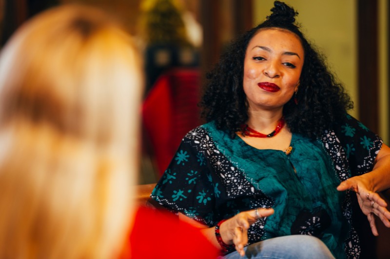 seated black woman gesticulating while chatting to another person in a dimly lit but cosy-looking indoor space