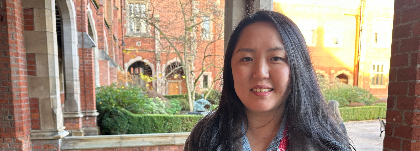 A woman with long dark hair stands smiling in an outdoor cloister area, with red-brick university buildings and gardens in the background.