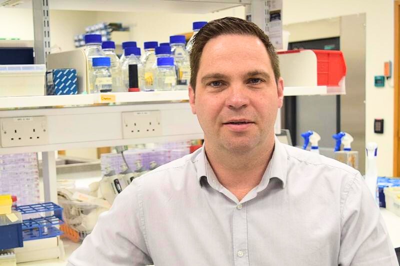 man wearing light-coloured shirt pictured in front of shelves in a laboratory space