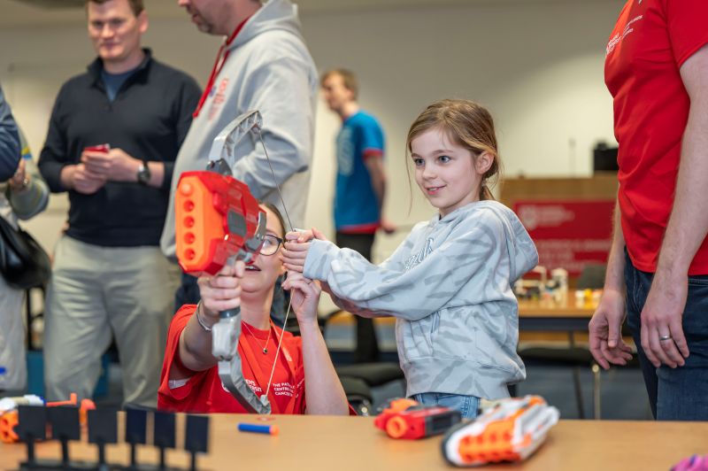 a smiling young girl aiming a Nerf-style bow and arrow with assistance, surrounded by other people milling around, at an indoor event