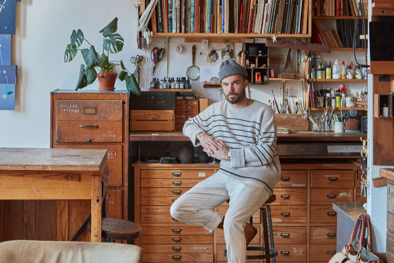 man wearing beanie hat looking at camera while sitting on a high stool in an airy art studio space with books, shelves, paints and worktops behind and around him