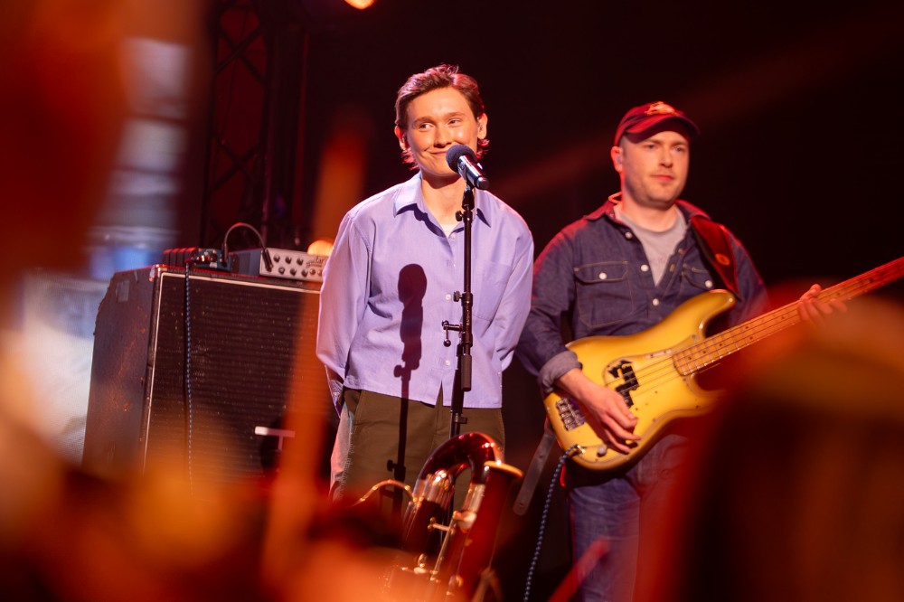 person wearing light blue shirt standing smiling behind a microphone stand with guitar player to their left