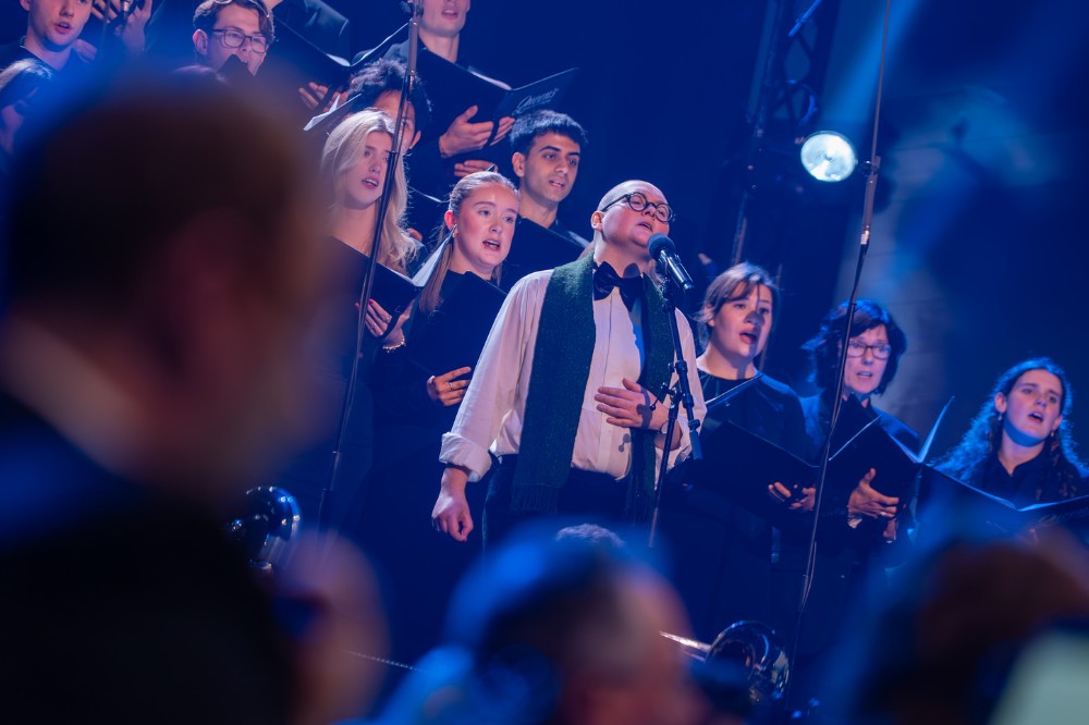 person singing on a blue-lit stage with a choir behind them and blurry people in the foreground