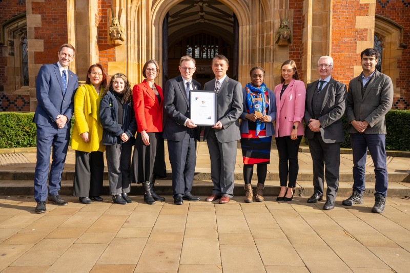 group of academics and professionals posing with a framed award certificate and trophy on the steps outside the front door of an old, redbrick building