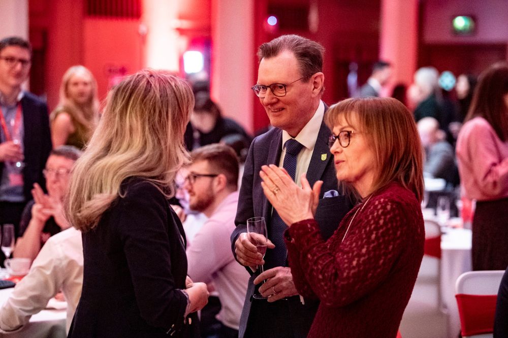 smiling besuited man wearing glasses and two smartly dressed women chatting over a sparkling drink at an indoor event