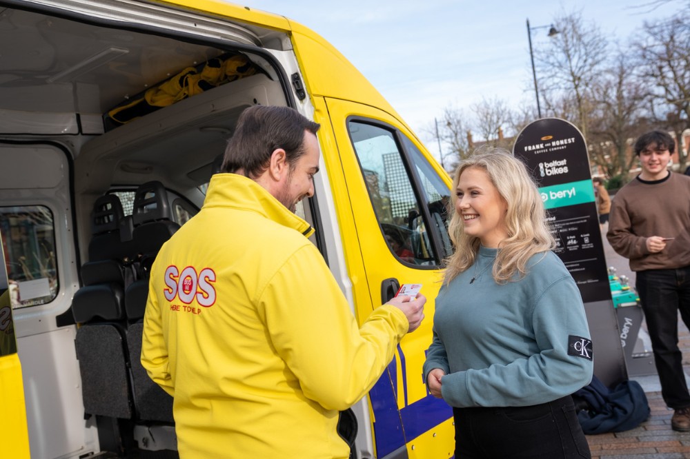 man in yellow jacket chatting to a young woman and checking an ID card beside a yellow minibus with people in the background