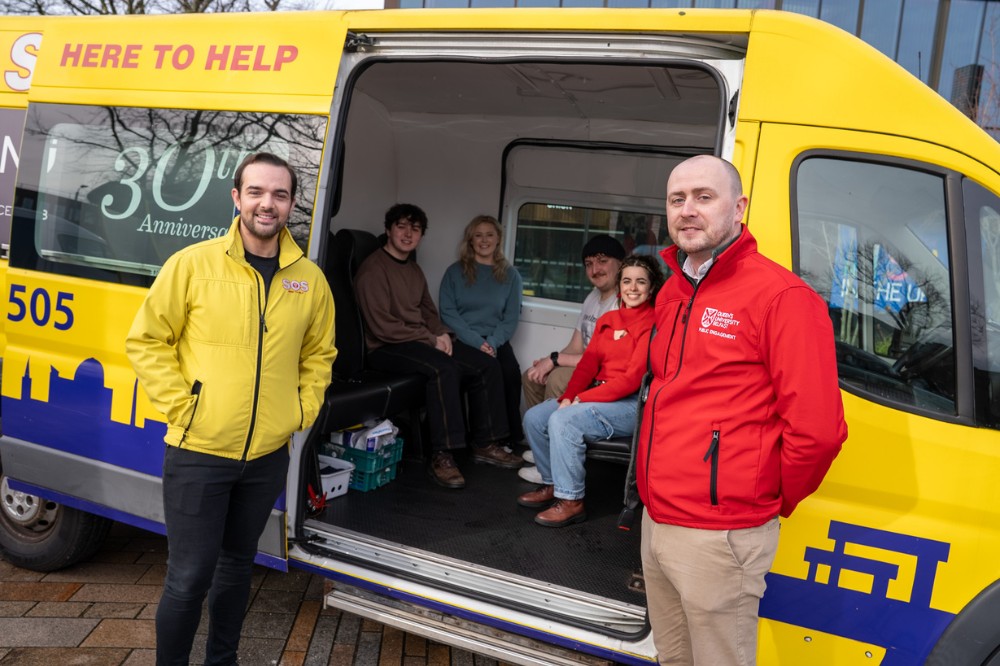 man in yellow jacket, left, and man in red zip-up sweater, right, framing the open door of a yellow minibus with smiling young people sitting inside