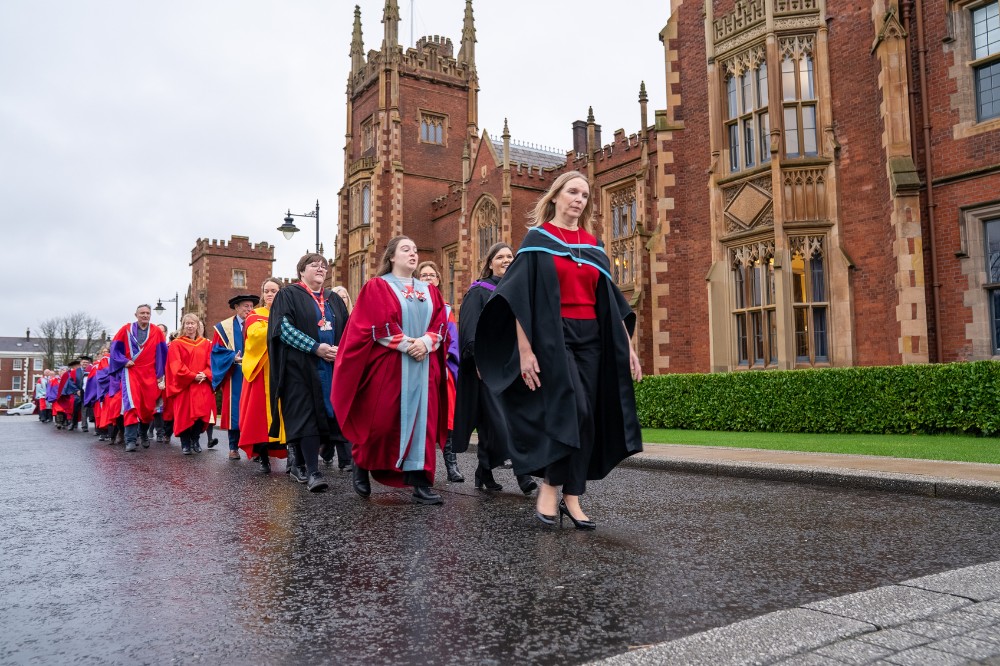 an academic procession, in graduation robes, processing in front of an old redbrick building